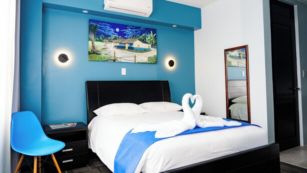 Een hotelkamer met een bed, nachtkastje, blauwe stoel, een luchtverfrisser aan de muur en een schilderij.