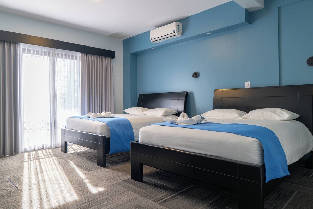 Hotellihuone, jossa on kaksi sänkyä, suuri ikkuna, jossa on verhot, ilmastointilaite ja matolla peitetty lattia.