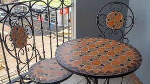 Een balkon met een metalen tafel en stoelen, uitzicht op een straat en bergen in de verte.