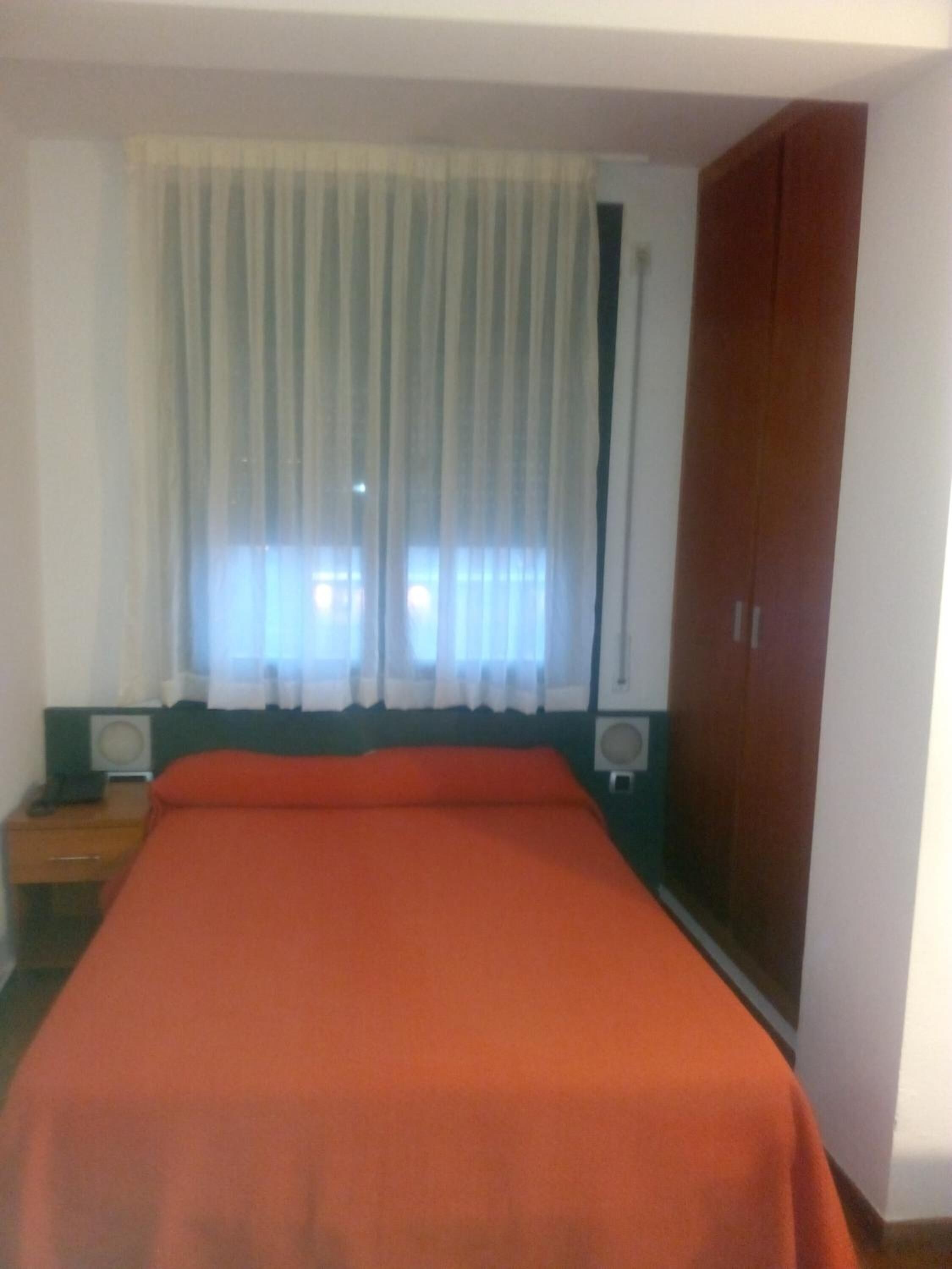 A room with a bed, a wardrobe, a window with curtains, and a bedside table with an alarm clock.