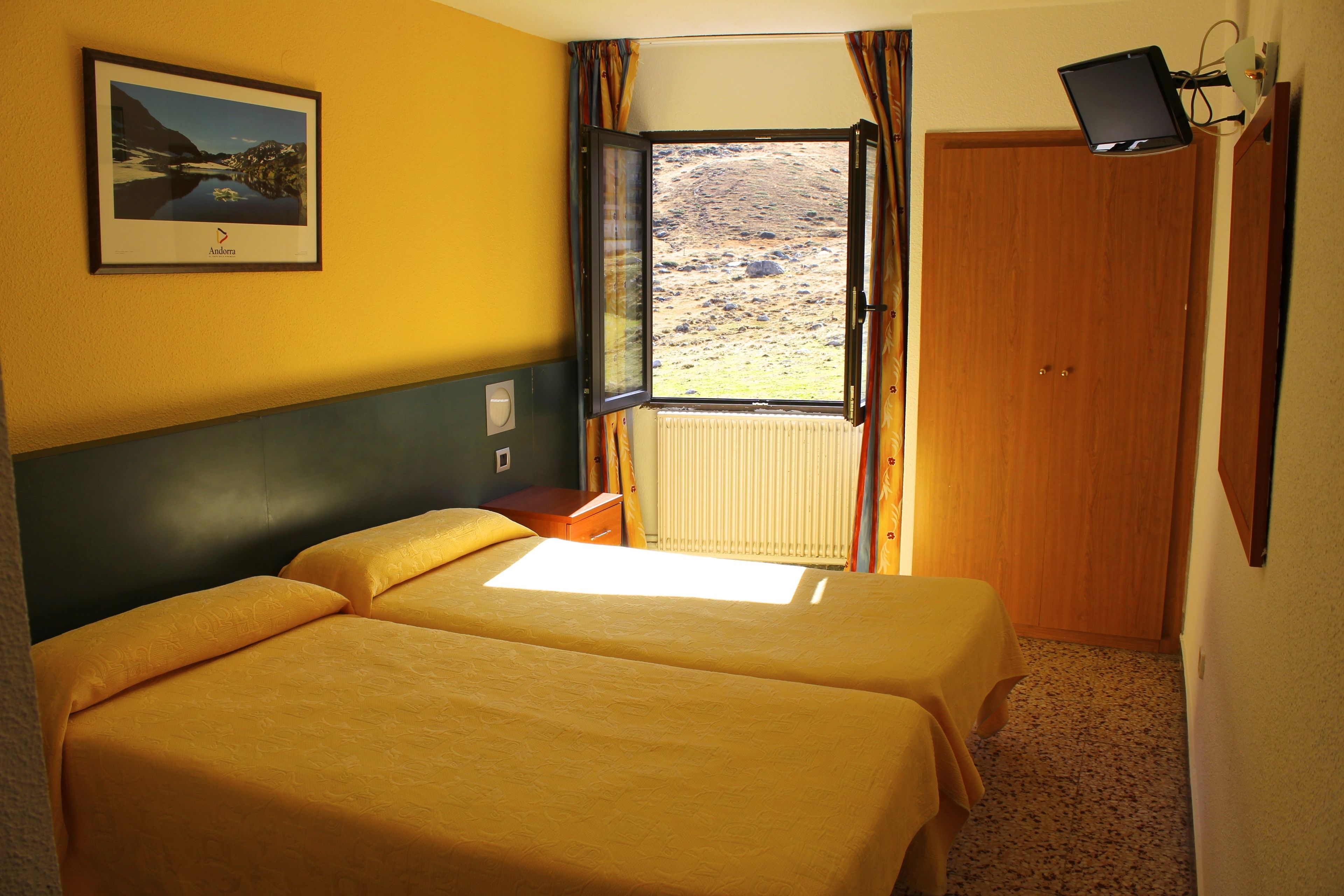 A room with two beds, a window with curtains, a framed picture on the wall, and a television mounted on the wall.