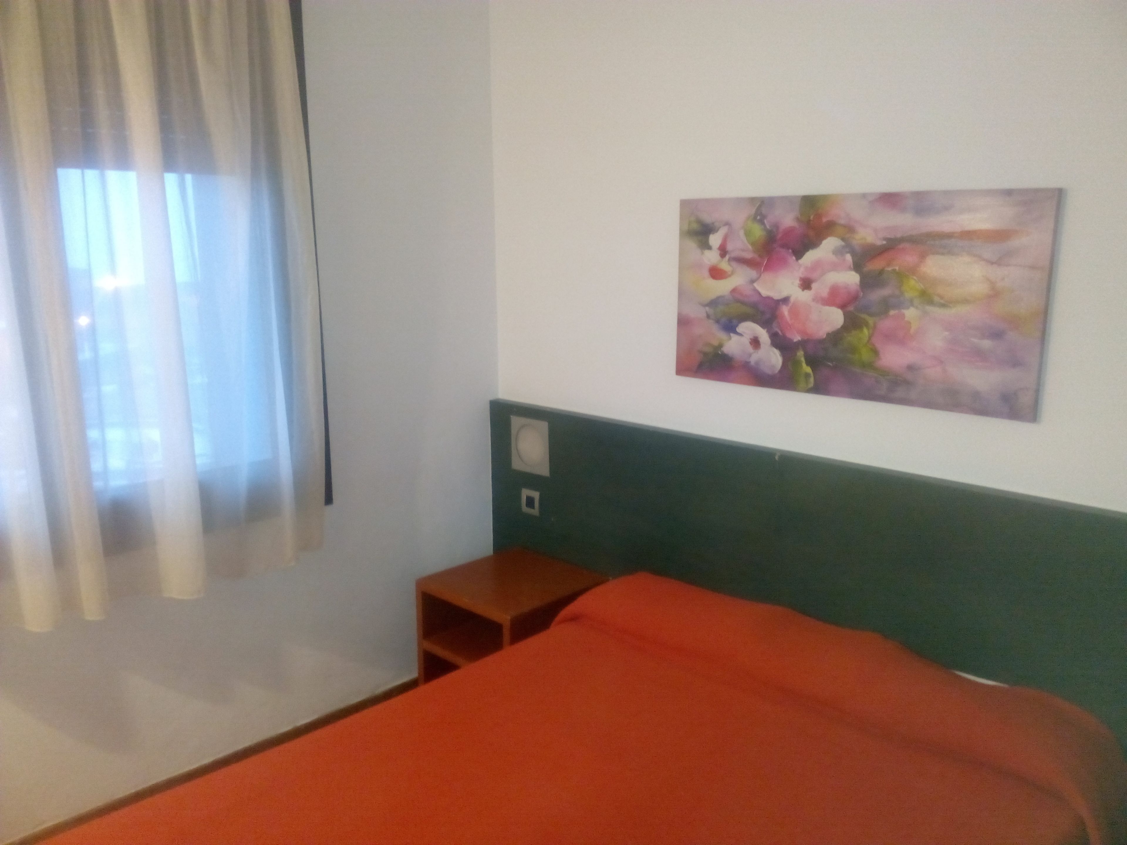 A hotel room with a bed, a small wooden nightstand, a painting on the wall, and a window with sheer curtains.