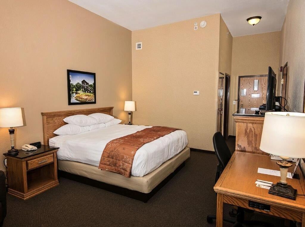 A hotel room with a bed, two bedside tables with lamps, a desk with a chair, a television, and a painting on the wall.