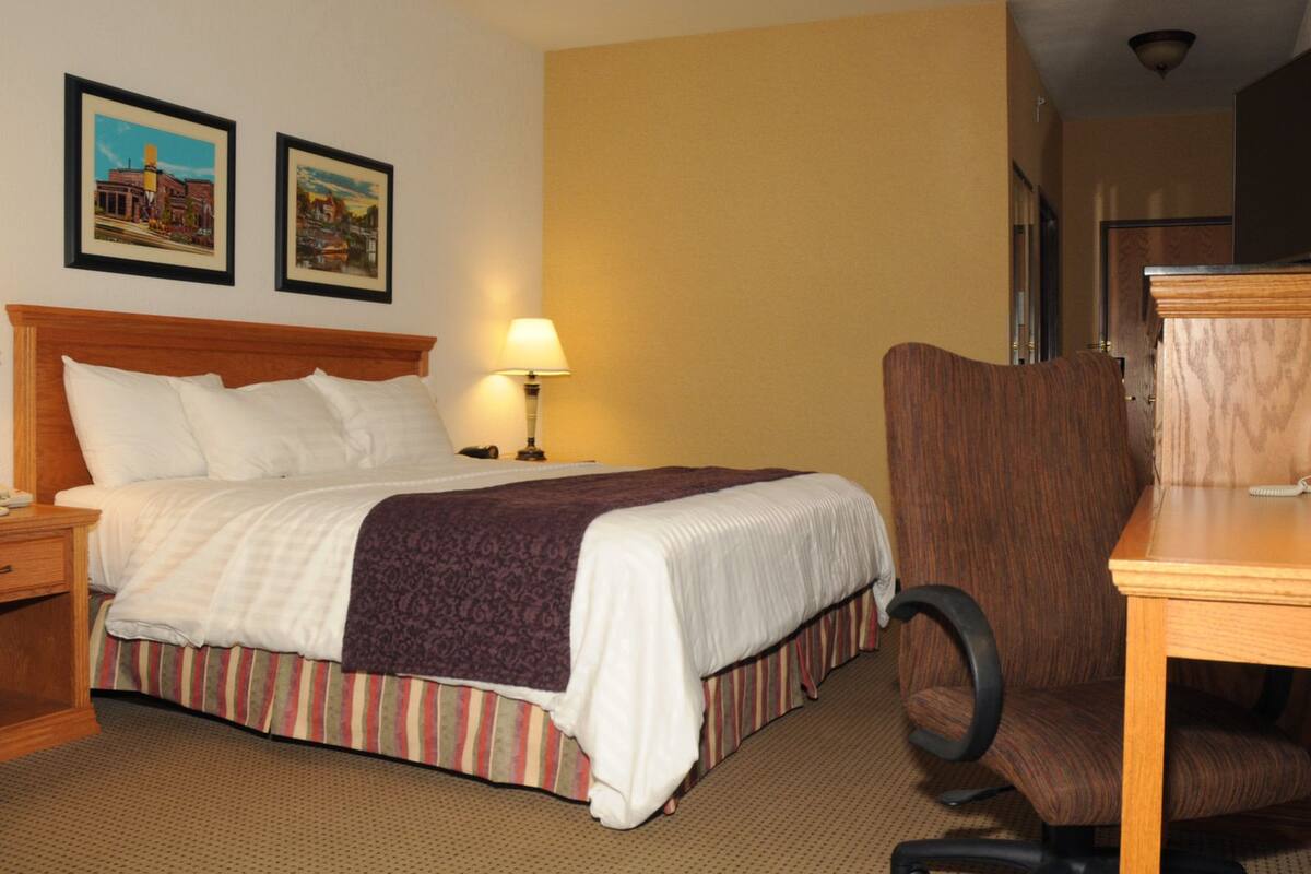 A hotel room with a bed, a desk, a chair, a lamp, and two framed pictures on the wall.