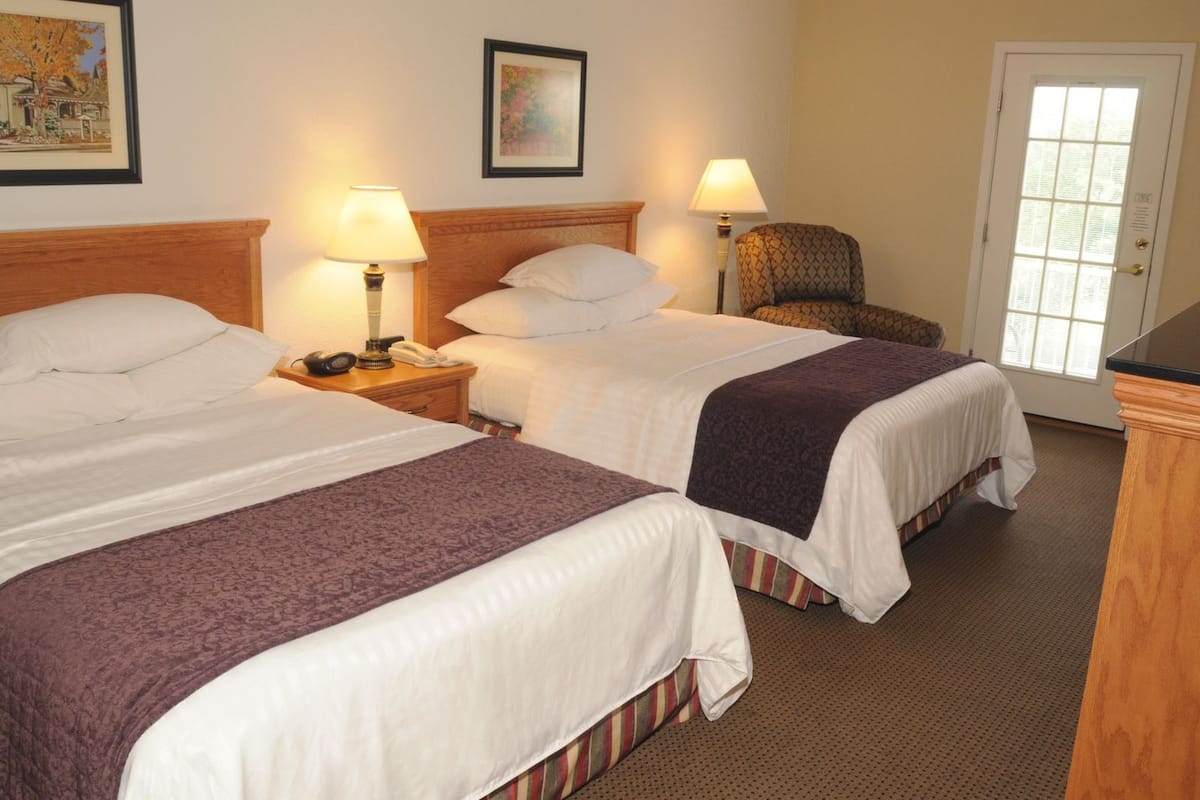 A hotel room with two beds, a wooden headboard, bedside lamps, a chair, and a door with a window.