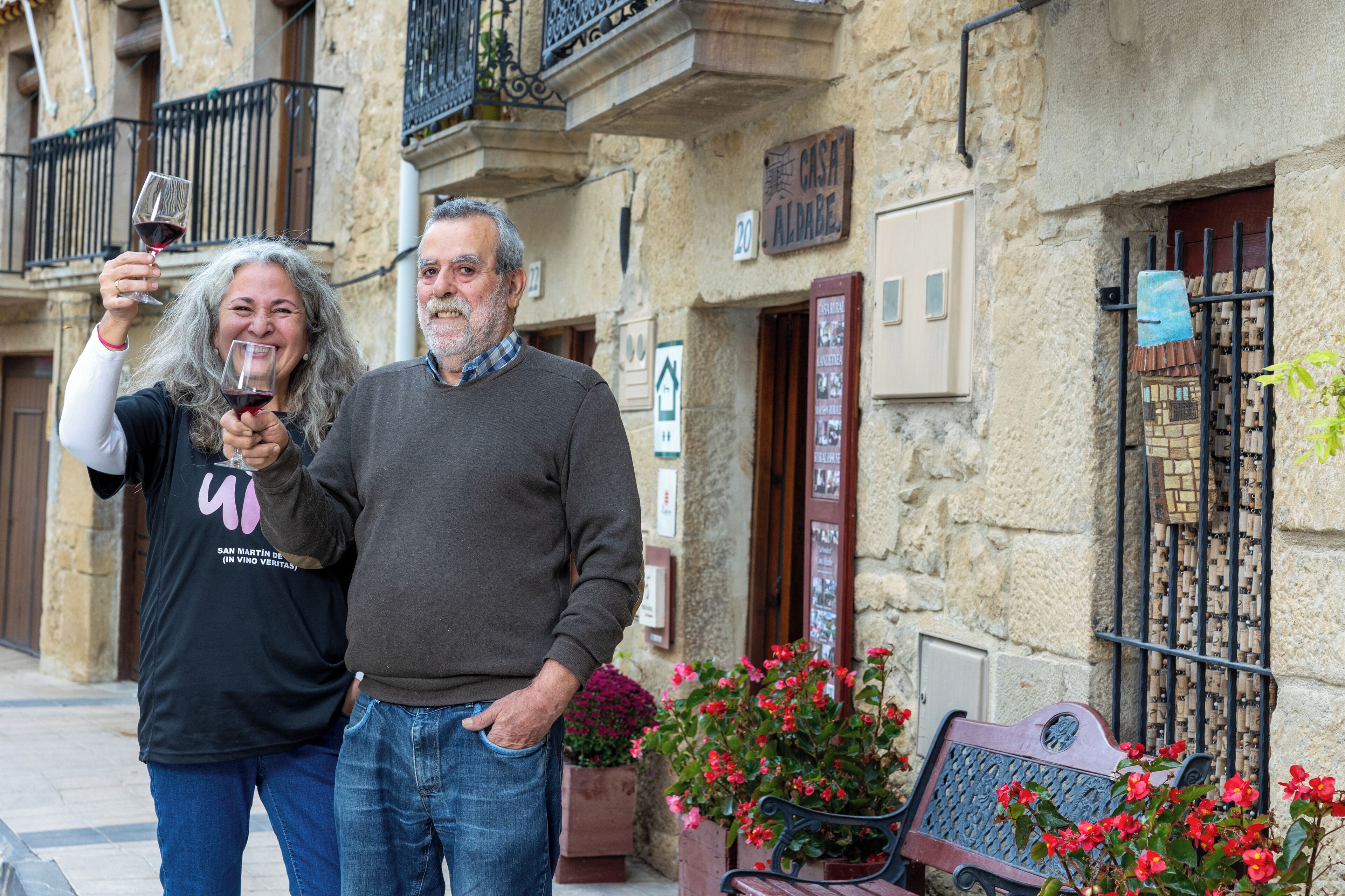 Un hombre y una mujer están de pie frente a un edificio de piedra con balcones, un banco y plantas en macetas.