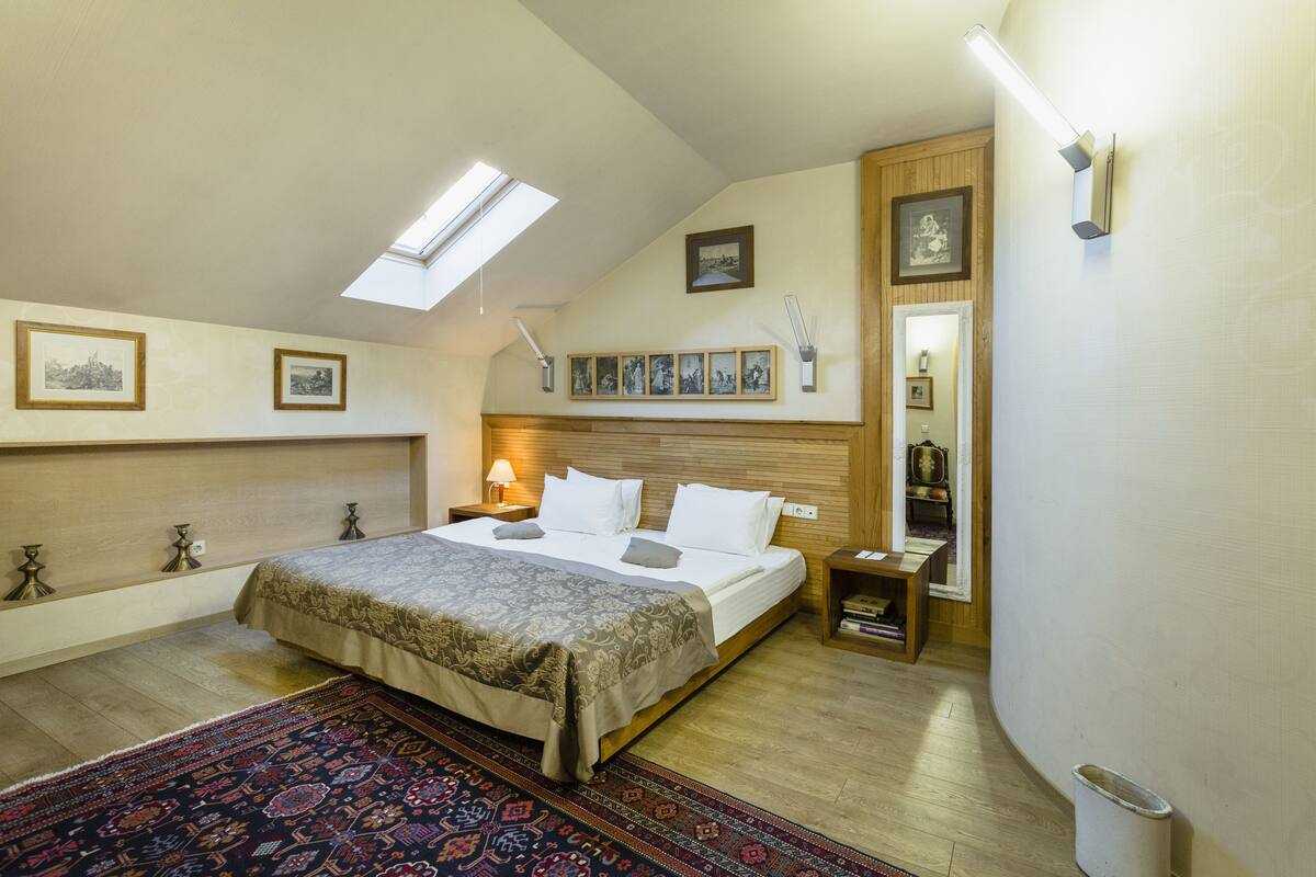 Un dormitorio con tragaluz, una cama con colcha estampada, una mesita de noche, un cabecero de madera y una estantería en la pared con cuadros.