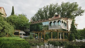Ein modernes Gebäude mit Glasfassade, einem Balkon mit Geländer und einem Garten mit Brunnen.