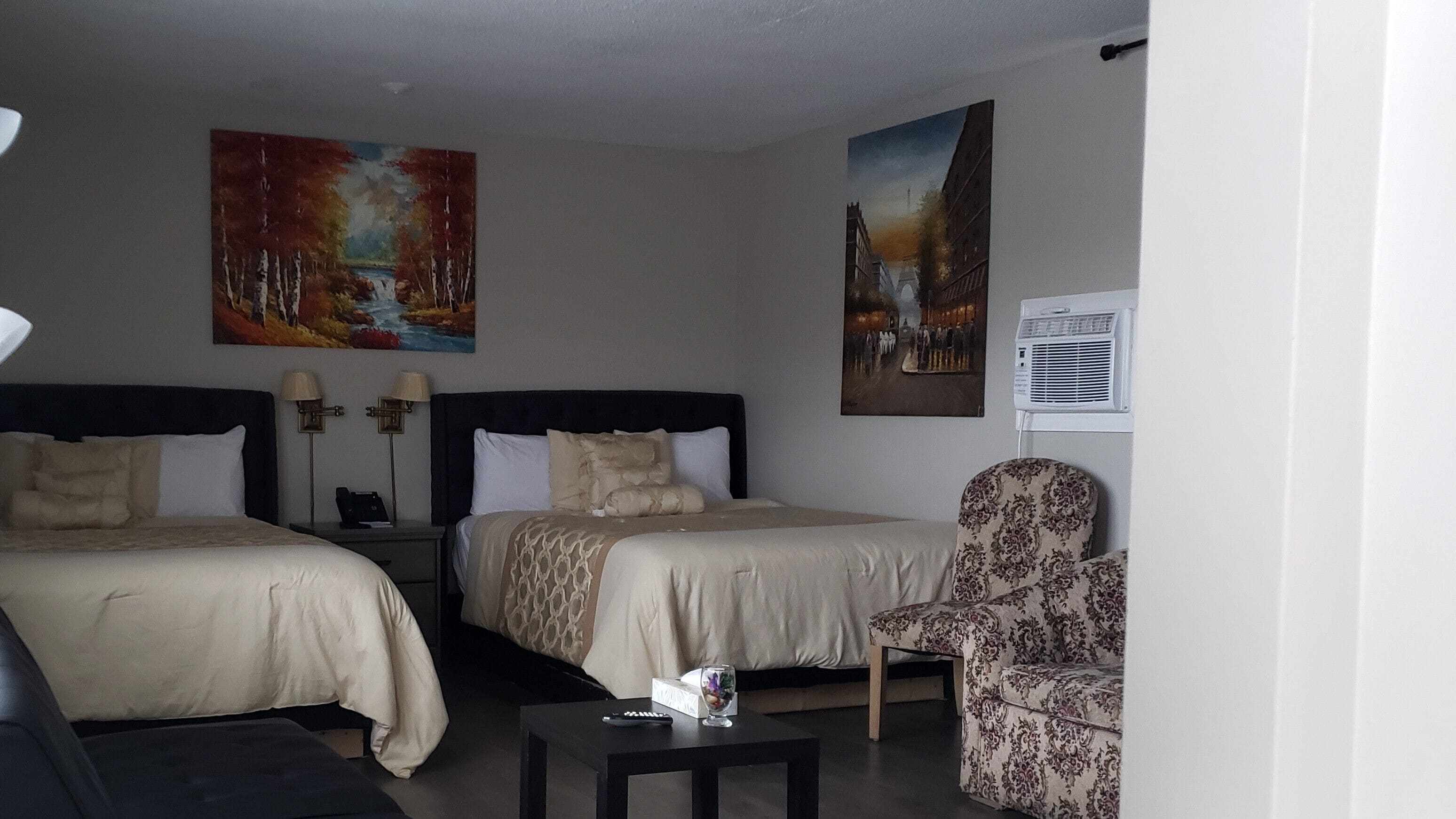 Una habitación de hotel con dos camas, una silla, una mesita y dos cuadros en la pared.