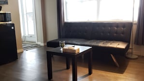 Una sala de estar con un sofá negro, una mesita con una caja y un vaso, un refrigerador negro y una ventana con cortinas.