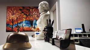 Una habitación con una puerta blanca, una escultura de busto, un cuadro de árboles otoñales y un escritorio con una campana, una taza negra y un calendario.