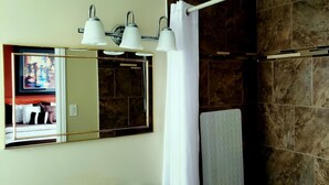 Un baño con un espejo grande, tres luces empotradas, zona de ducha con azulejos marrones y una toalla blanca colgada en un perchero.