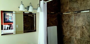 A bathroom with a large mirror, three wall-mounted lights, a shower area with brown tiles, and a white towel hanging on a rack.