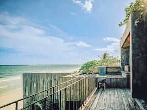 A wooden deck with a railing overlooking a beach, a concrete structure with greenery, and a clear view of the ocean.