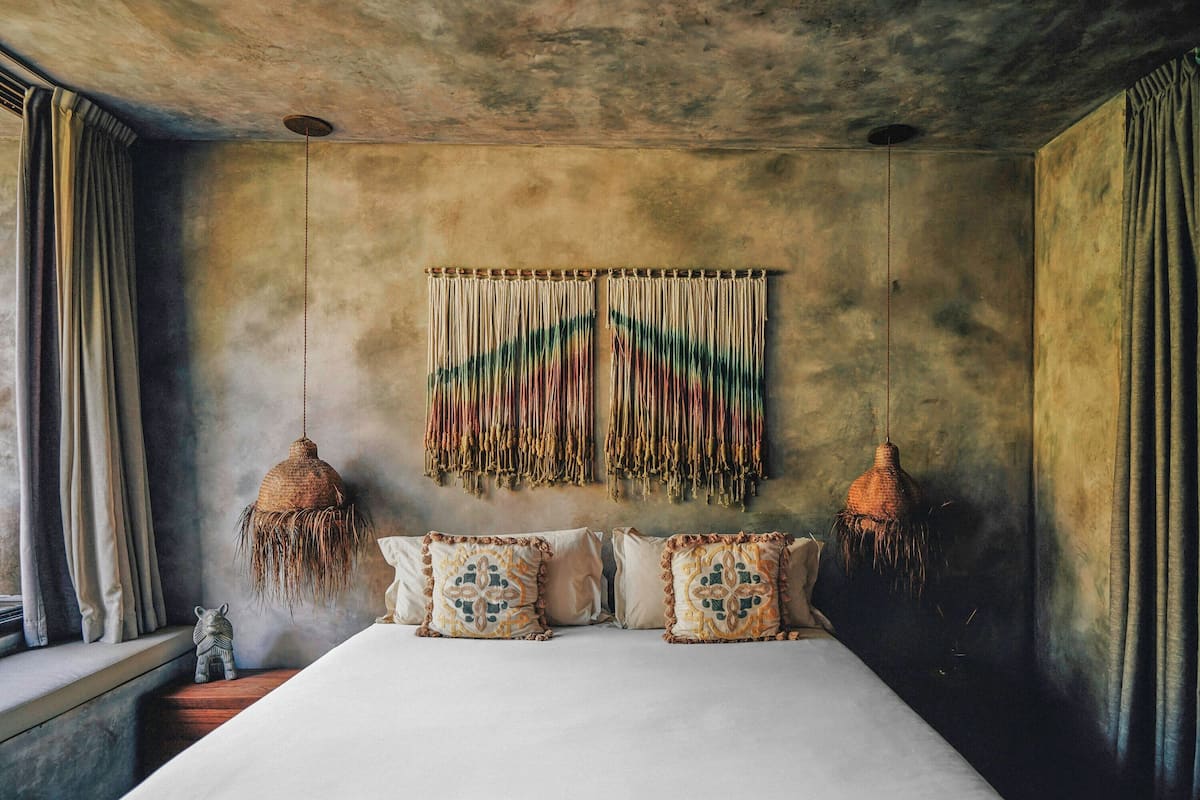 Una cama con almohadones estampados, un colgador de macramé y dos lámparas colgantes.