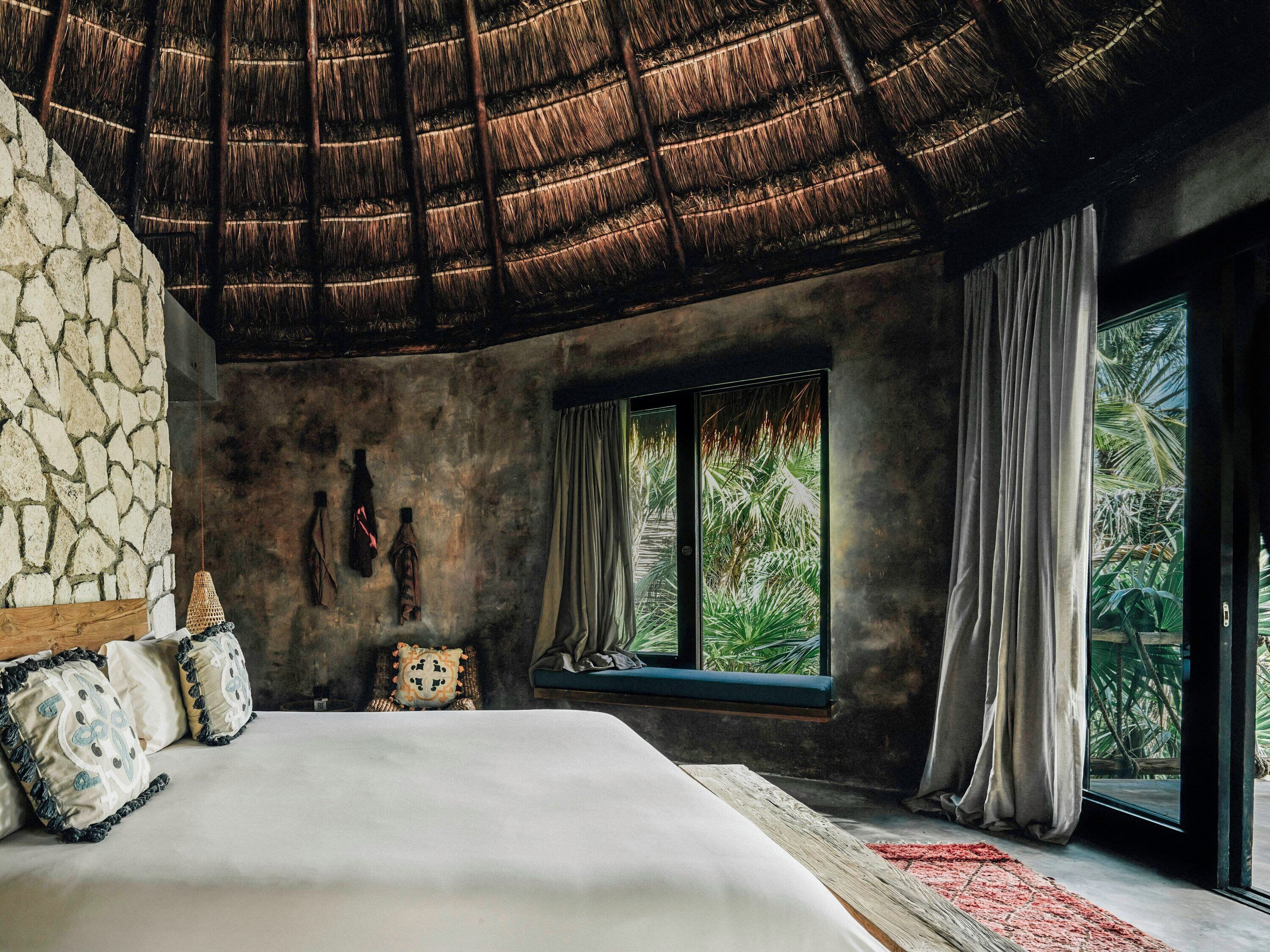 A bedroom with a large bed, stone walls, a thatched ceiling, and a window with curtains.