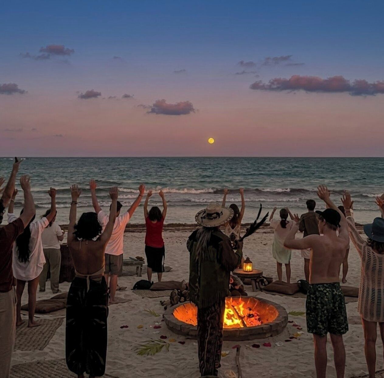 A group of people at a beach with a fire pit in the foreground.