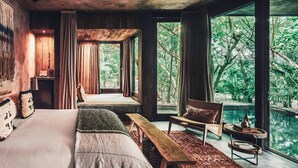 Un dormitorio con una cama grande, un banco, una silla y vistas a un paisaje natural a través de amplias ventanas.