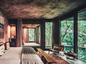 A bedroom with a large bed, a bench, a chair, and a view of a natural landscape through large windows. - Nômade Temple Tulum (Tulum)