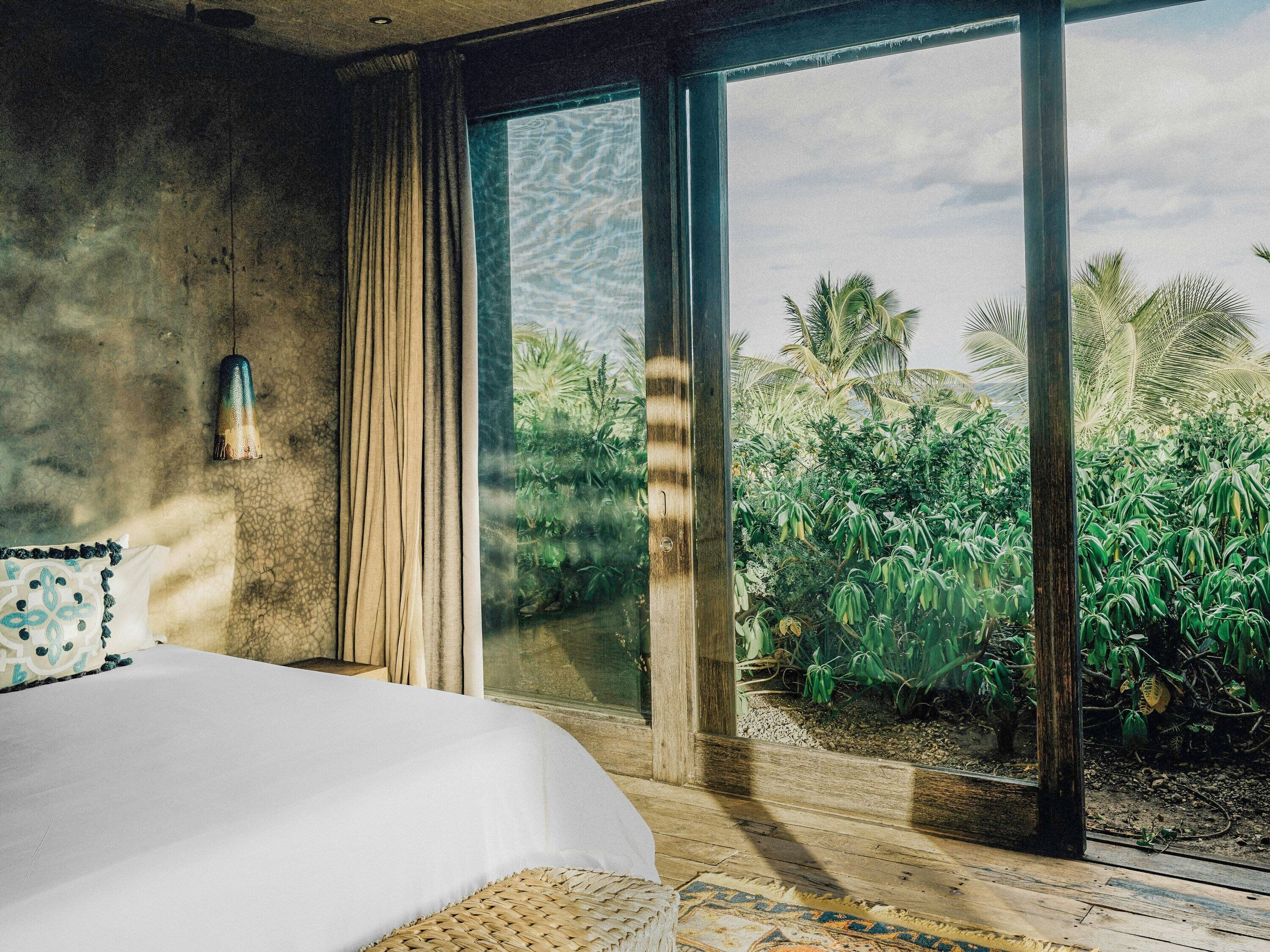 Une chambre avec un grand lit, une vue sur la végétation tropicale par une porte vitrée et un mur en béton.