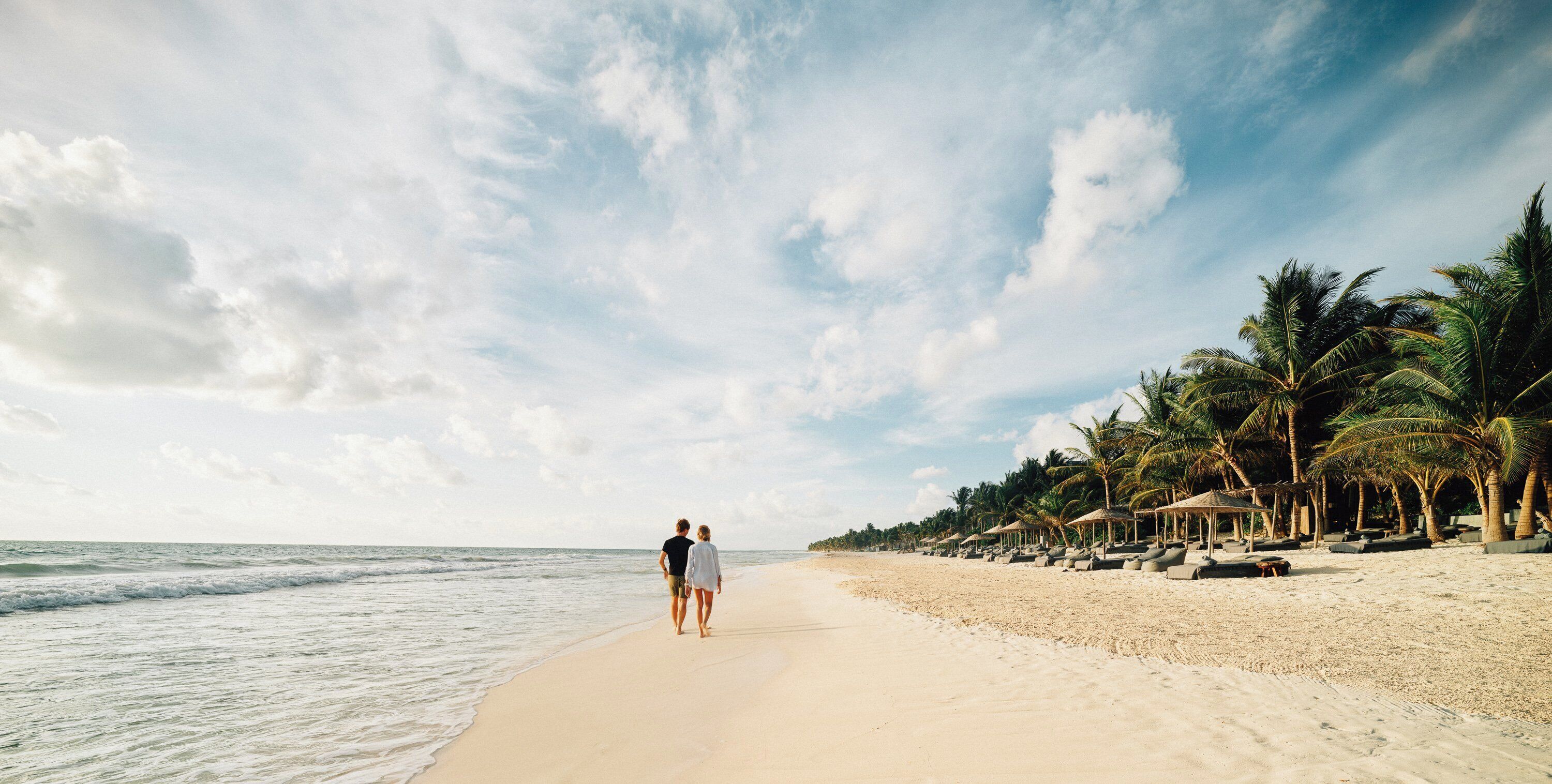 A beach with a couple walking along the shore, palm trees, and beach huts.