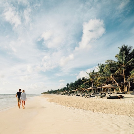 A beach with a couple walking along the shore, palm trees, and beach huts.