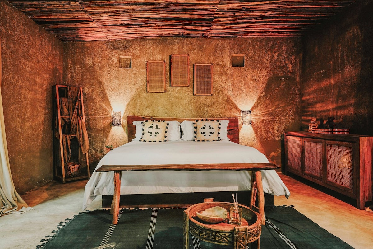 Un dormitorio rústico con una cama de madera, una silla tejida y una mesita con un cuenco y una vela.