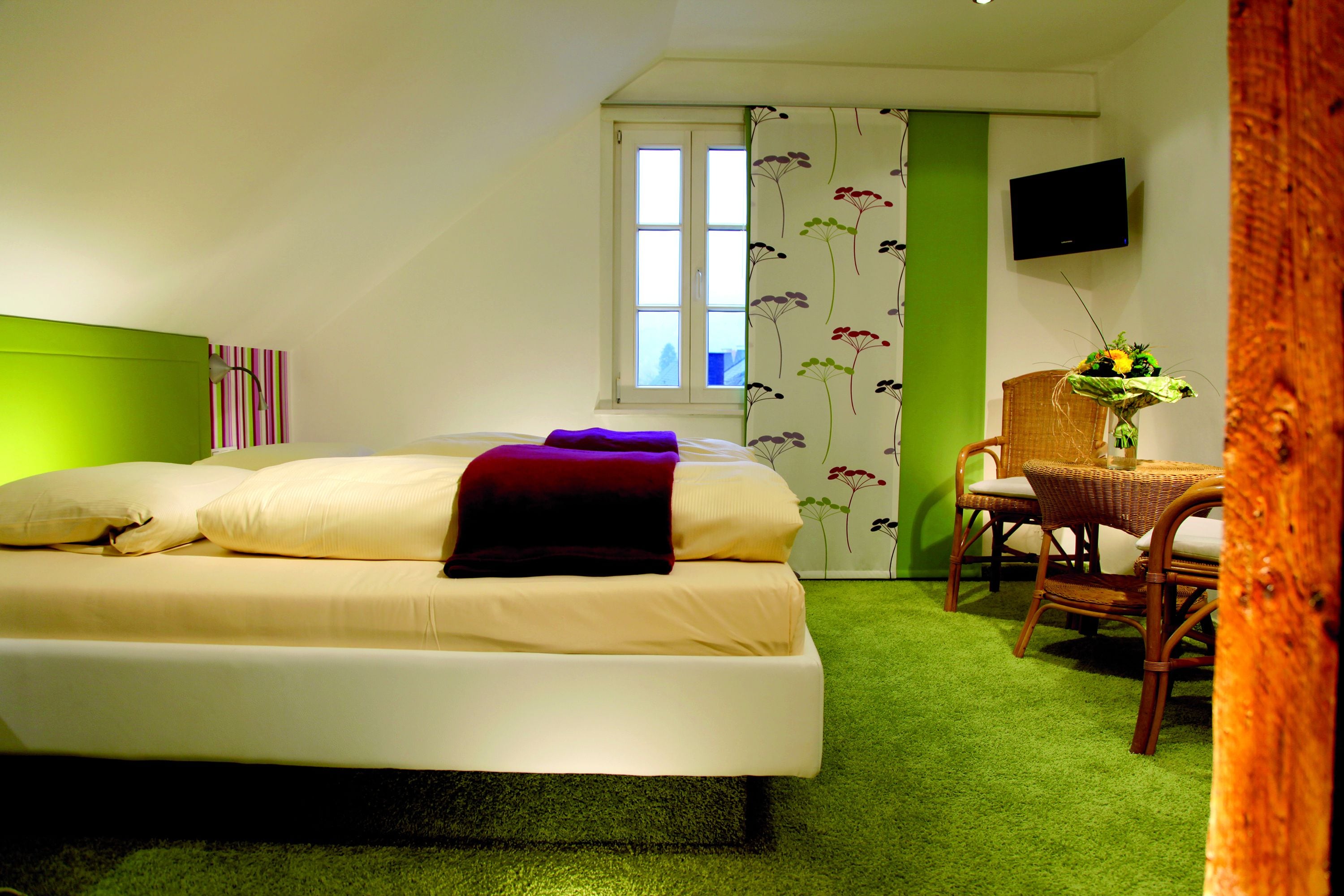 Un dormitorio con cama, una silla, una mesa con un florero, una ventana y un televisor de pantalla plana.