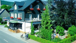 A large, dark-colored hotel with multiple windows and balconies, surrounded by lush greenery and a clear blue sky. - Flair Hotel Nieder (Bestwig)