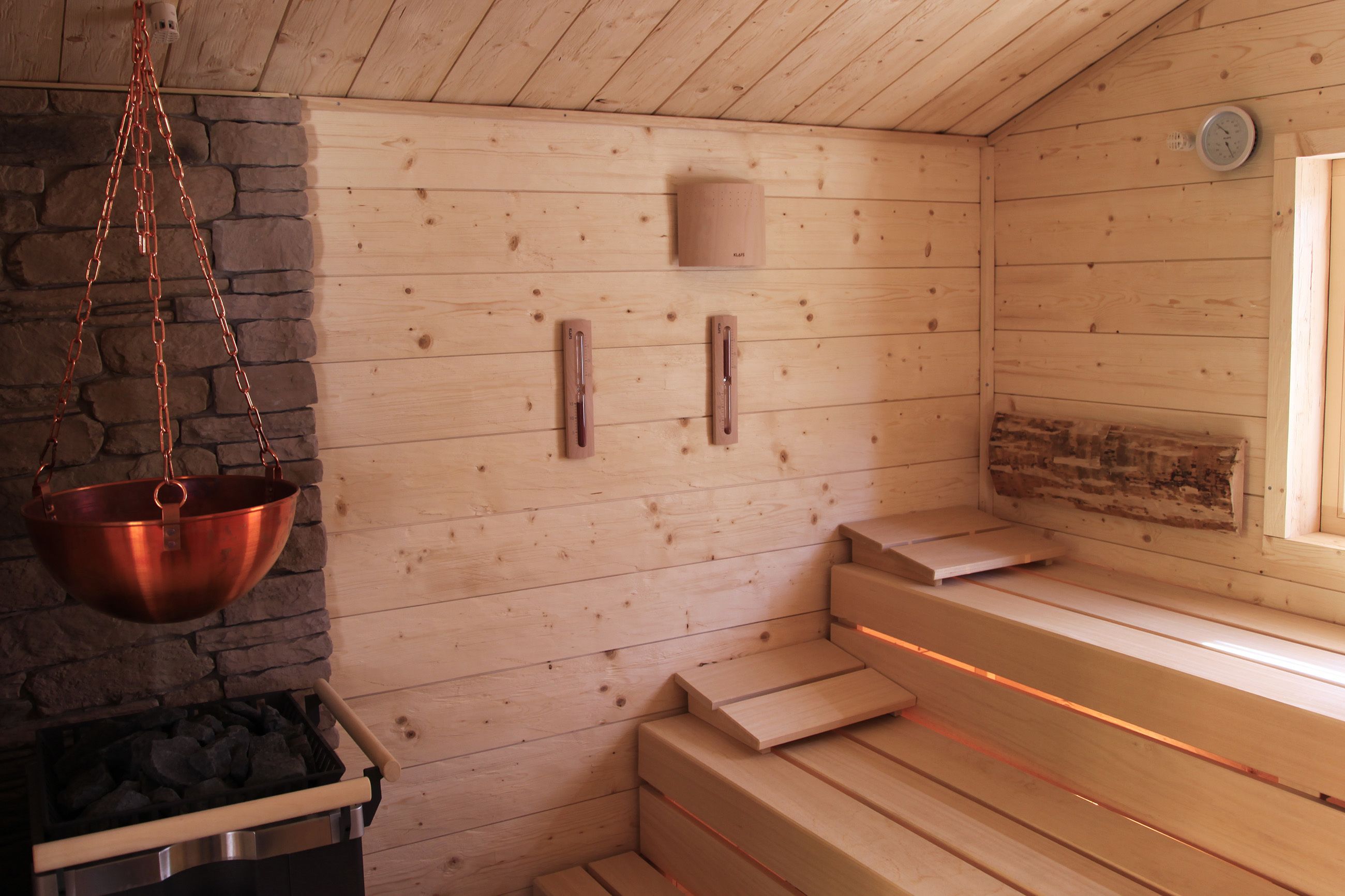 Una sauna con bancos de madera, una pared de piedra y un recipiente de cobre.