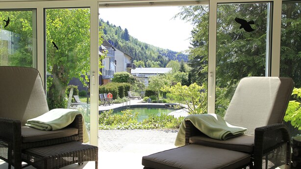 A room with two wicker chairs facing a glass door, a small pool, and a view of a house and forested hill. - Flair Hotel Nieder (Bestwig)