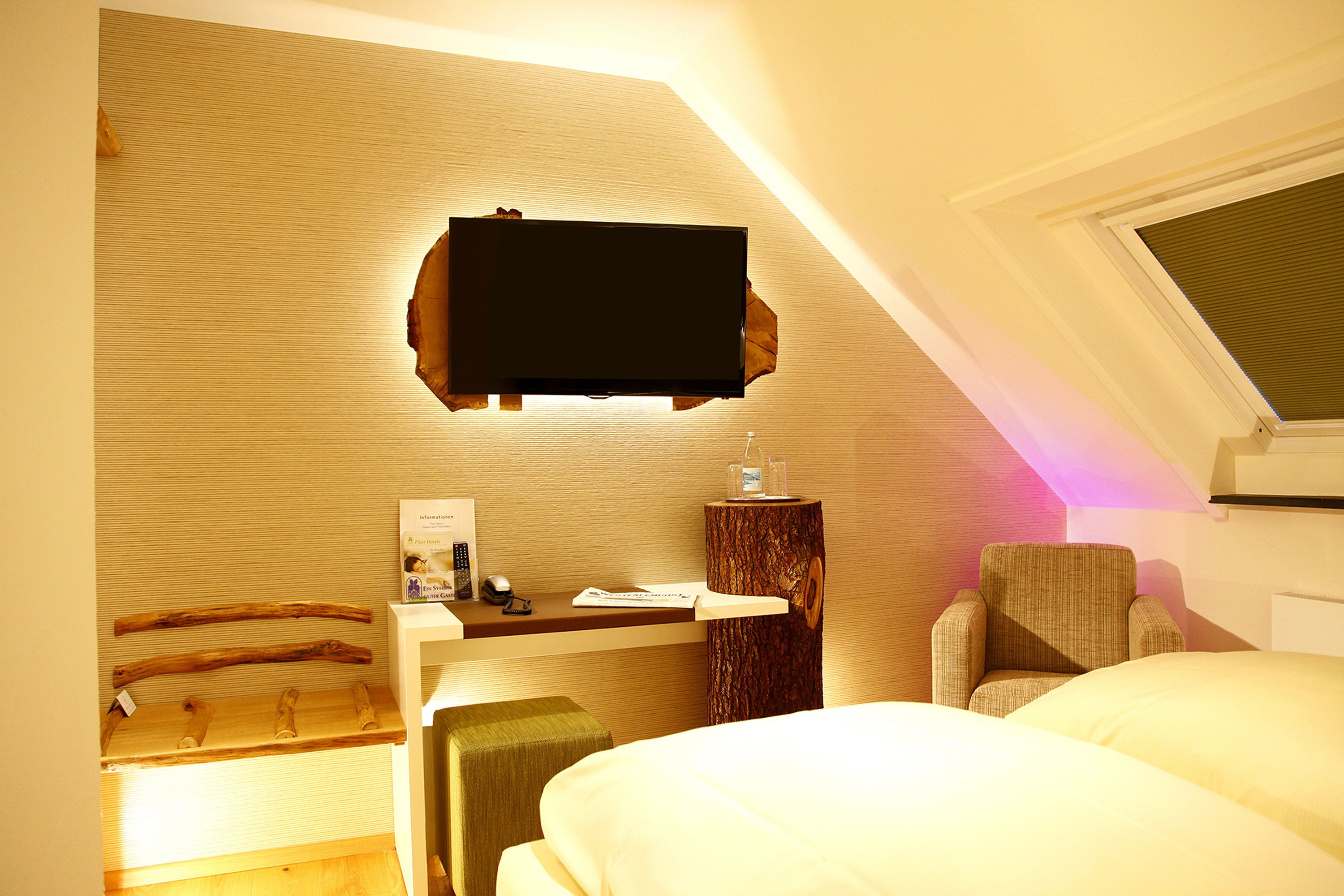 Habitación de hotel con televisor de pantalla plana en la pared, un escritorio de madera con silla, una mesita de noche y una cama.