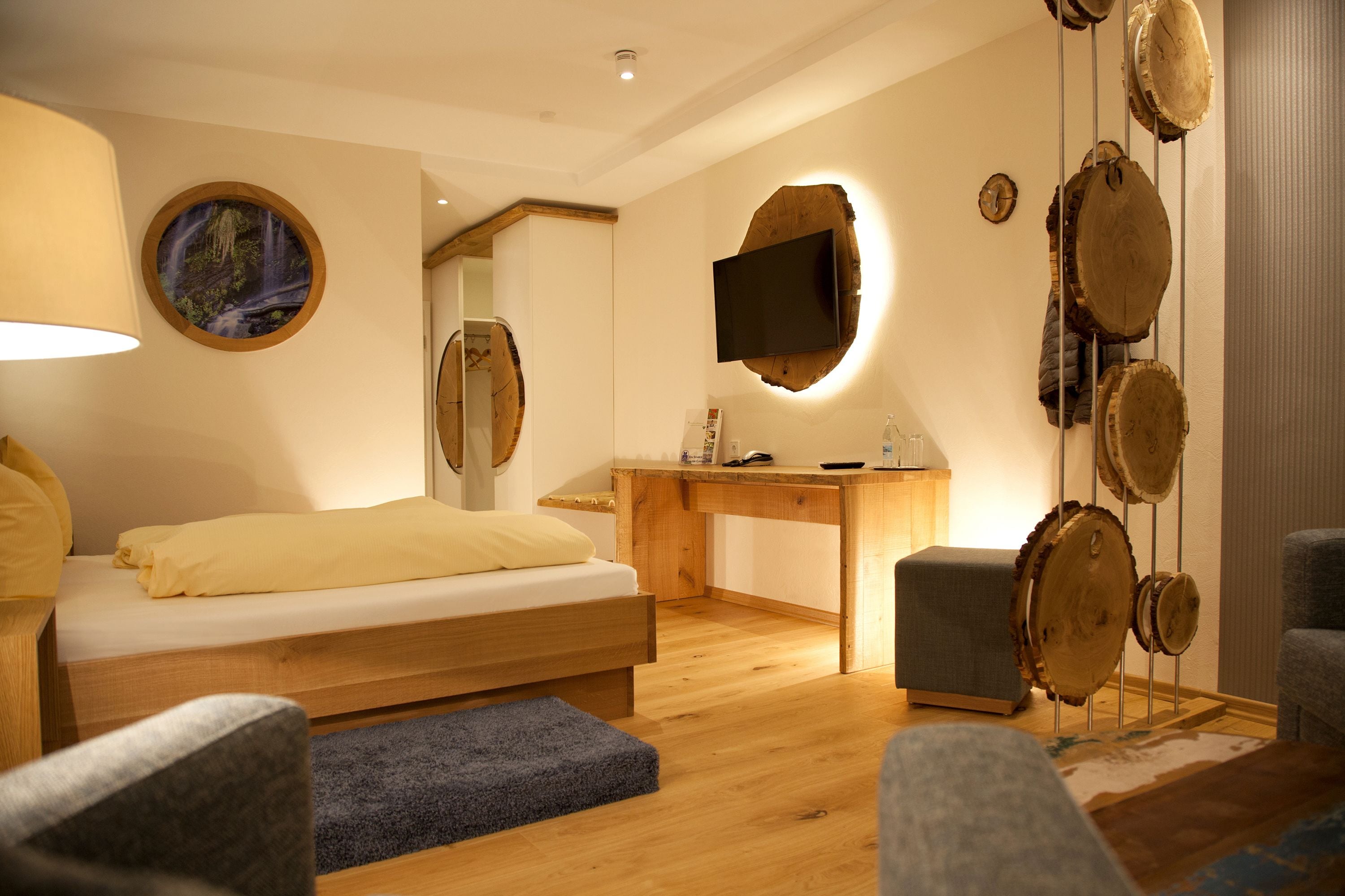 A modern bedroom with a wooden bed, a desk, a TV mounted on a tree trunk, and a unique wall decoration of wooden discs.