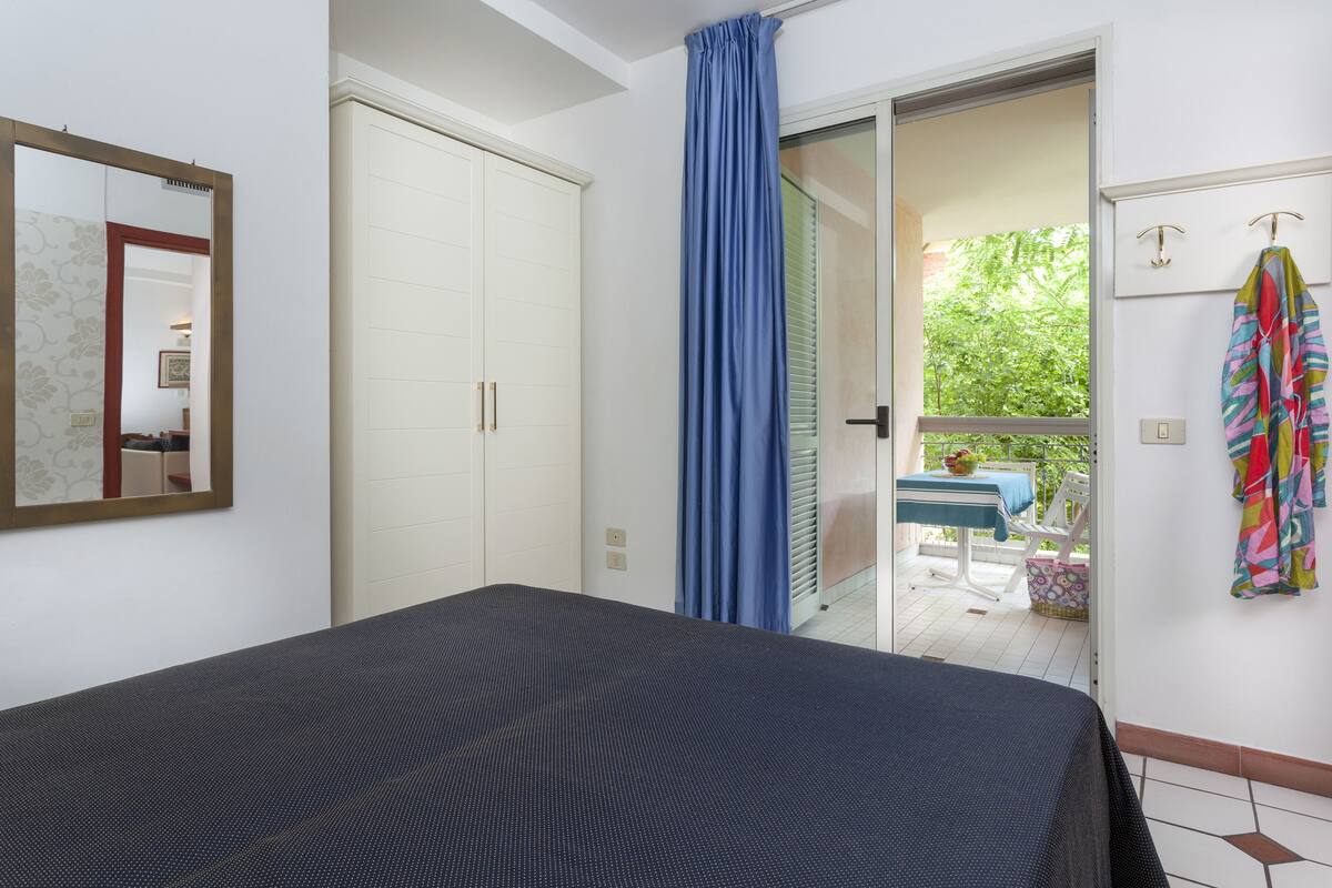 A bedroom with a bed, a wardrobe, a bathroom door, and a view of an outdoor area with a table and chairs.