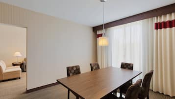 Habitación de hotel con una mesa de comedor de madera, cuatro sillas estampadas, un sofá, una lámpara y un ventanal con cortinas.