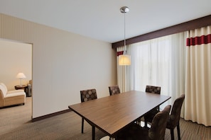 Una habitació d'hotel amb una taula de dinar de fusta, quatre cadires estampades, un sofà, una làmpada i una finestra amb cortines.