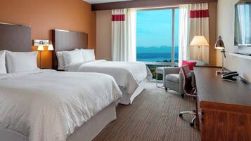 Een hotelkamer met twee bedden, een bureau, een stoel, een lamp en een groot raam met uitzicht op bergen.