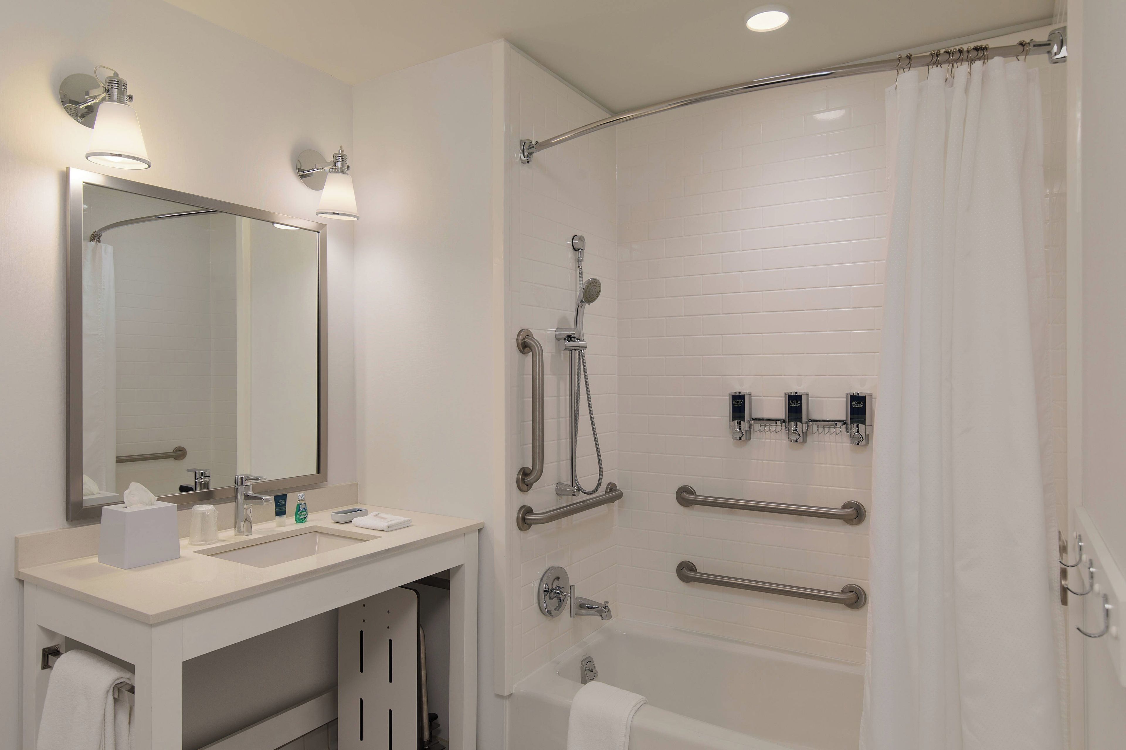 Une salle de bain moderne avec un meuble-lavabo blanc, un grand miroir, une douche équipée de barres d’appui et une baignoire.