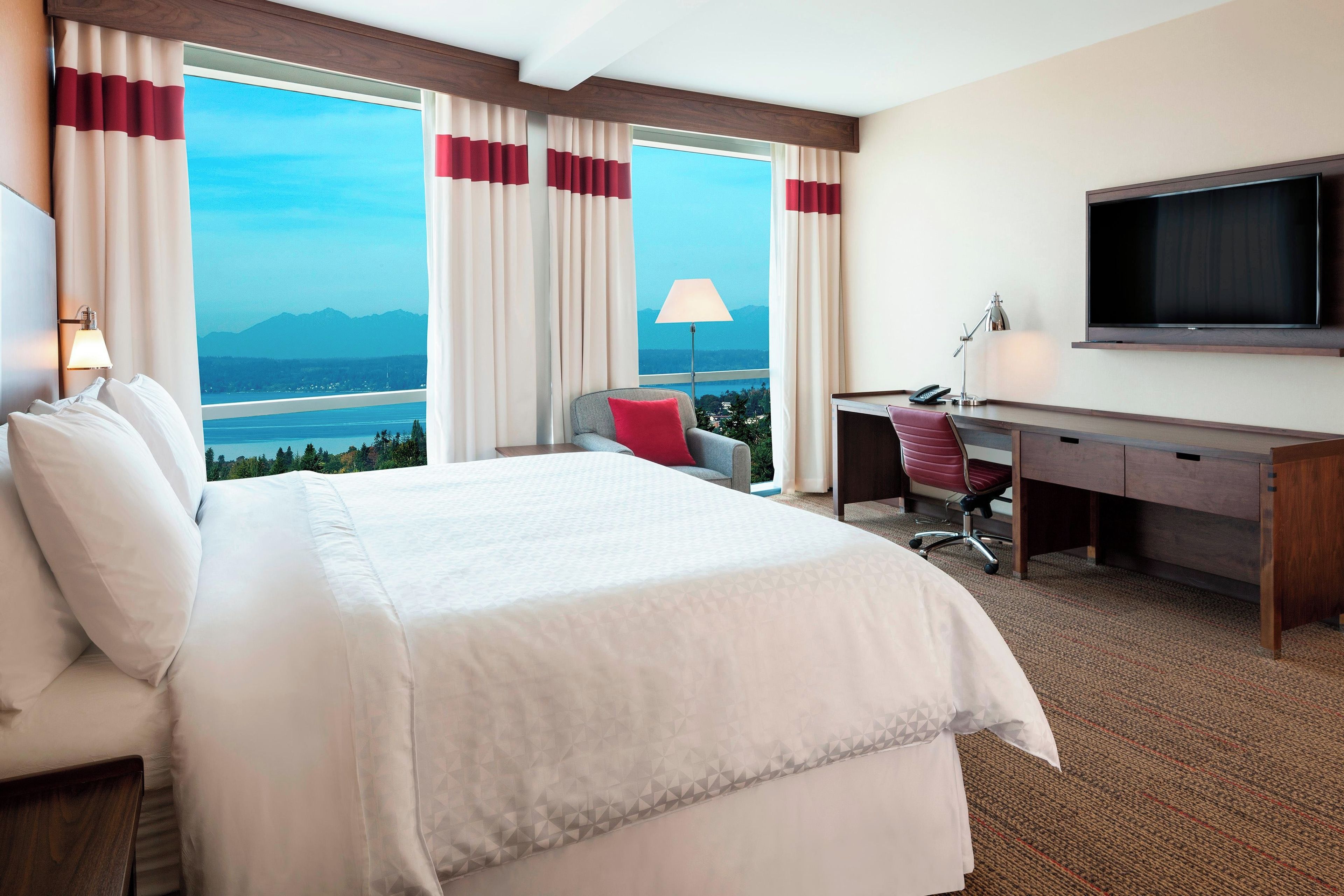 Habitación de hotel con cama, escritorio, silla, televisor y vista a un cuerpo de agua y montañas.