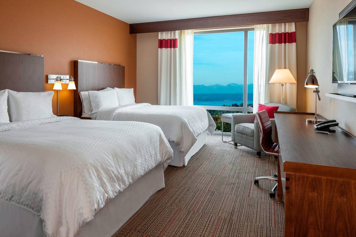 Une chambre d’hôtel avec deux lits, un bureau, une chaise, une lampe et une grande fenêtre donnant sur les montagnes.