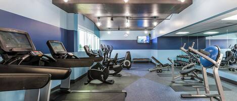 A gym with various exercise equipment, a mirrored wall, and a television screen.