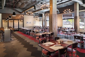 Een ruim restaurant met meerdere tafels en stoelen, grote ramen en hangende pendantlampen.