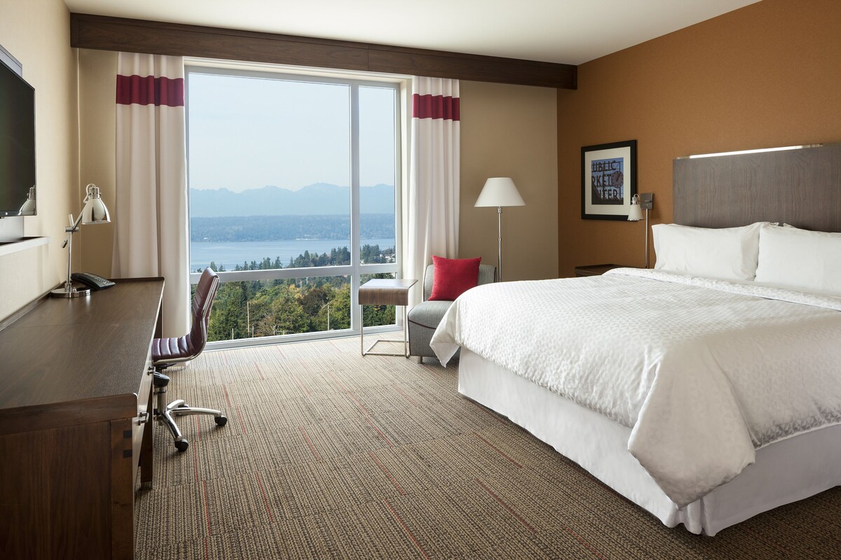 Une chambre d’hôtel avec un grand lit, un bureau et une chaise. La fenêtre offre une vue sur des montagnes et un plan d’eau.