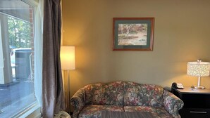 Une pièce avec un canapé à motifs floraux, une petite table, une lampe, un tableau encadré au mur et une fenêtre avec des rideaux.