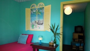 Una habitación con paredes verde azuladas, una cama roja, una mesa de madera con una lámpara, una planta en maceta y un cuadro de una escena tropical.