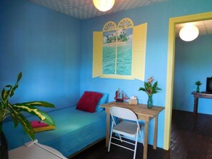 A room with a blue wall, a wooden desk, a white chair, a small table with a plant, and a window with a tropical scene. - Valeri Emanuel Apartments (Bocas del Toro)