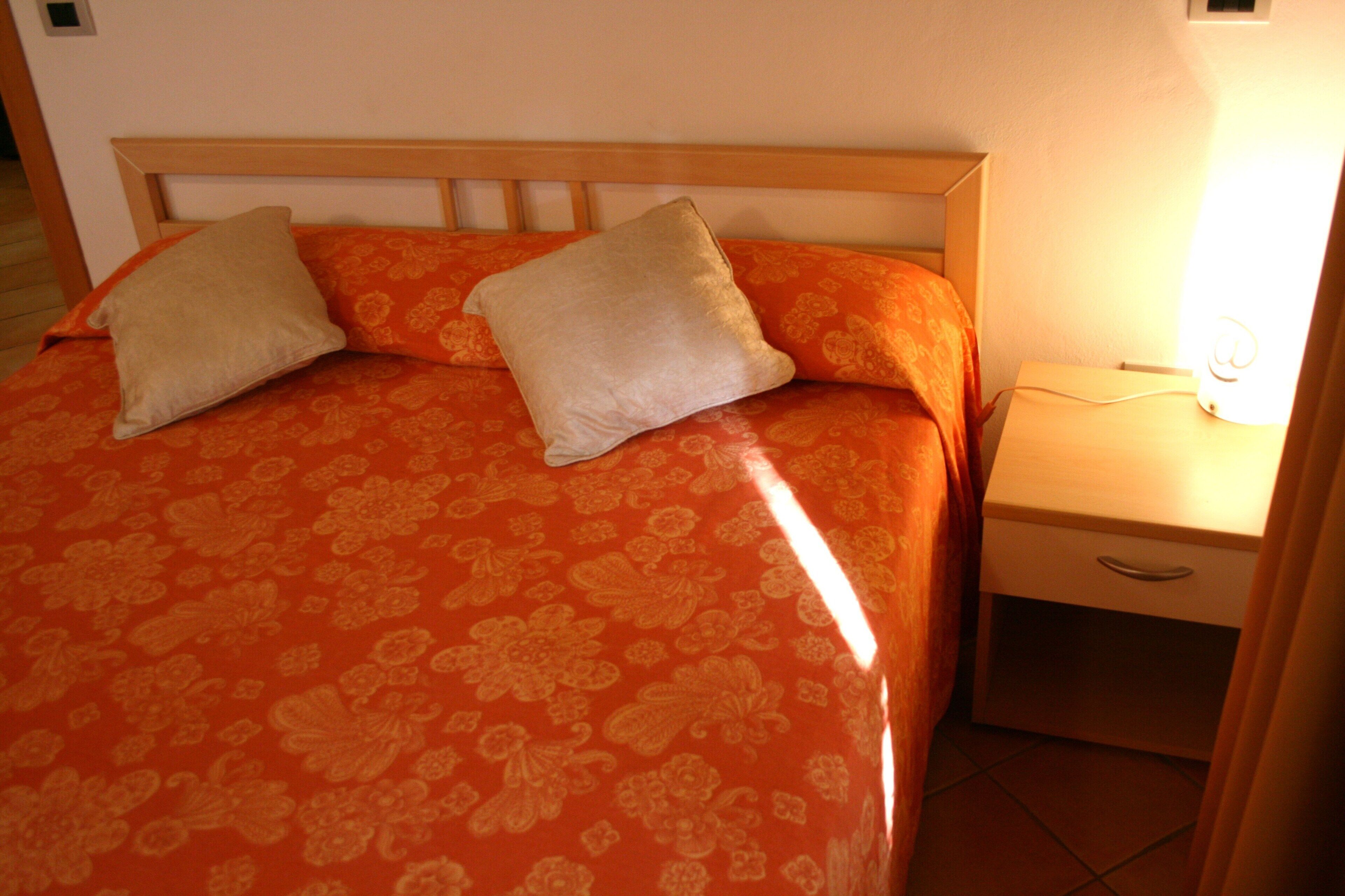A bed with a floral-patterned bedspread and two pillows, a bedside table with a lamp, and a nightstand with a drawer.