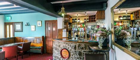 Uma área de bar com balcão de pedra, cadeiras de madeira e uma variedade de garrafas e copos nas prateleiras.