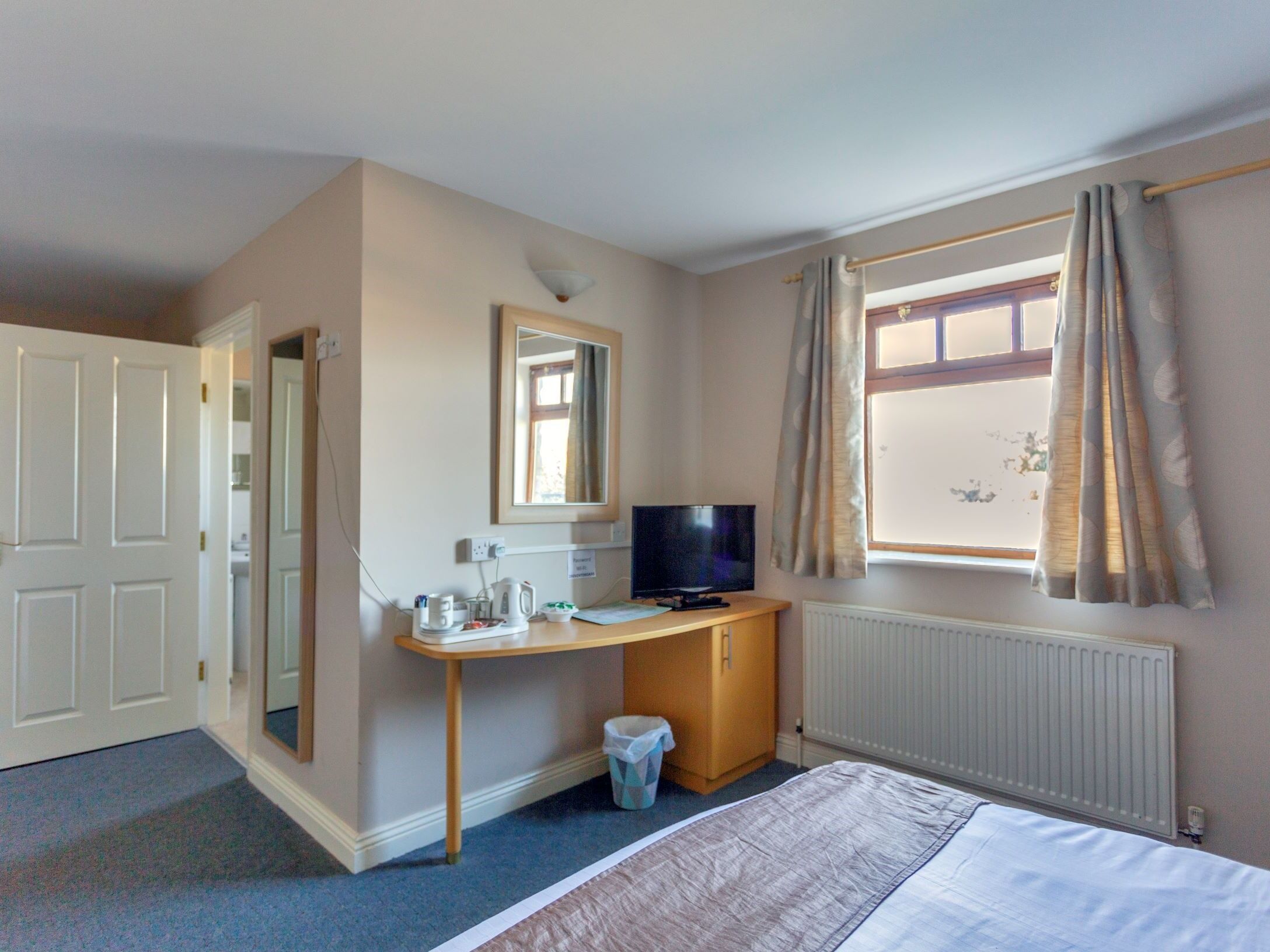 Une chambre d’hôtel avec un lit, un bureau avec une télévision, un miroir, un radiateur et une fenêtre avec des rideaux.