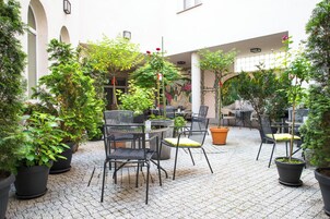 Un patio al aire libre con plantas en macetas, mobiliario exterior y vista a un edificio con arcos.
